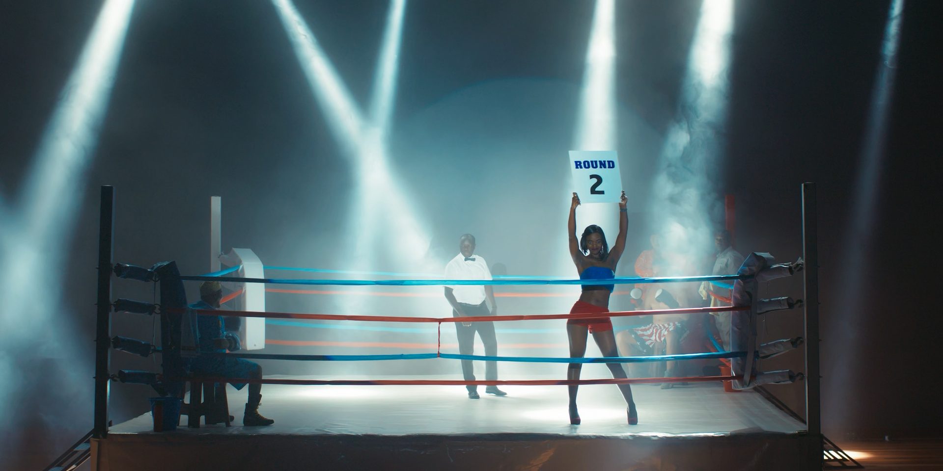 A boxing ring under spotlights, a woman holding up the Round 2 card