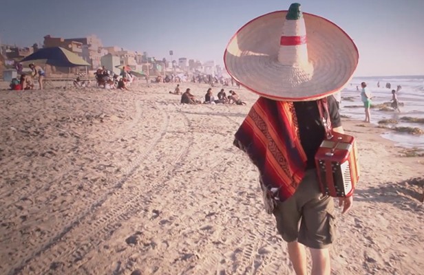 A man in a sombrero carries an accordion along a beach